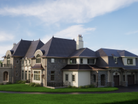 Exterior view of a grand custom estate designed by Ballard+Mensua Architecture, blending European-inspired detailing with stone and stucco cladding, steeply pitched slate roofs, copper accents, and arched dormers. Set against a manicured lawn and blue sky, the home reflects the firm’s expertise in high-end residential architecture with classical proportions and rich material palettes.