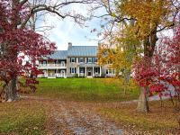 Traditional stone farmhouse with metal roof and symmetrical façade, featuring double porches with white columns and rocking chairs, set on a grassy hill framed by vibrant autumn trees.
