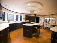 Contemporary custom kitchen with curved dark wood cabinetry, white solid-surface countertops, and a sculptural ceiling hood detail above a central island cooktop, designed by Ballard+Mensua. Warm wood floors and integrated dining area highlight craftsmanship and modern family living.