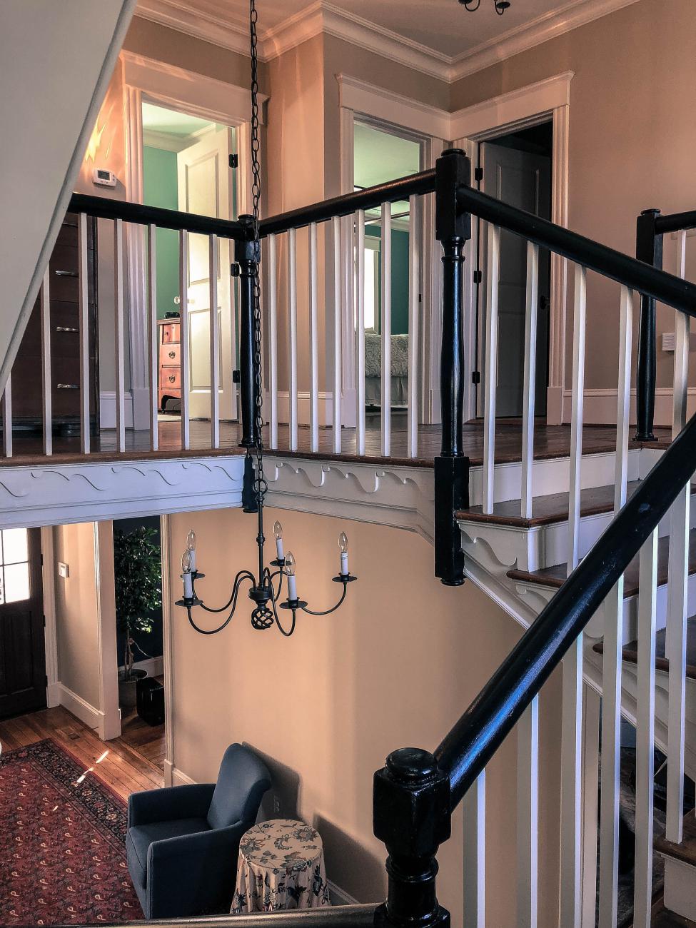 Two-story stair hall with classic wood balustrade, black handrails, and decorative trim, featuring a central iron chandelier and warm-toned walls, leading to bedrooms with painted walls and natural light.