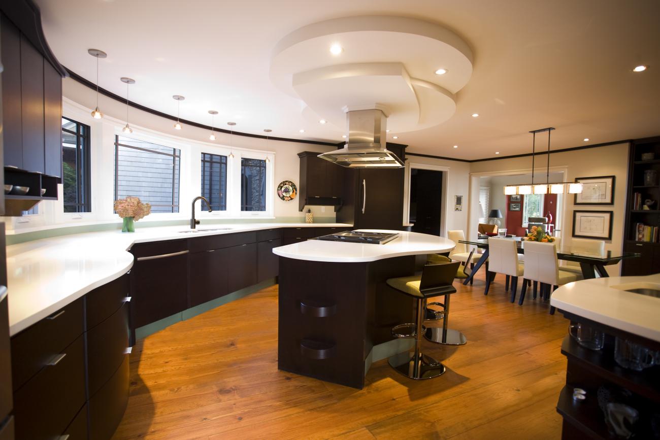 Contemporary custom kitchen with curved dark wood cabinetry, white solid-surface countertops, and a sculptural ceiling hood detail above a central island cooktop, designed by Ballard+Mensua. Warm wood floors and integrated dining area highlight craftsmanship and modern family living.